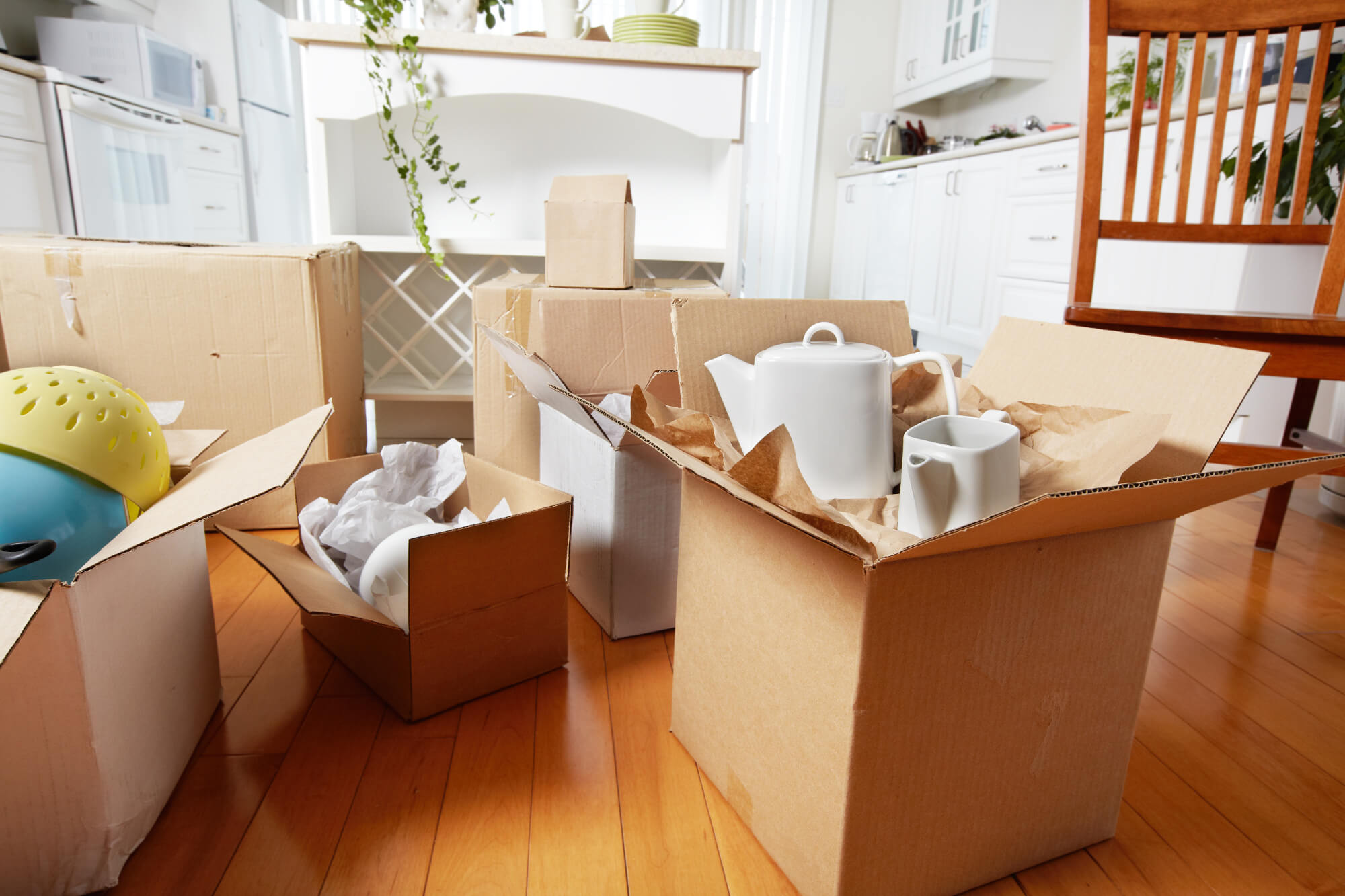 Open cardboard boxes with household items on a wooden floor inside a bright kitchen.