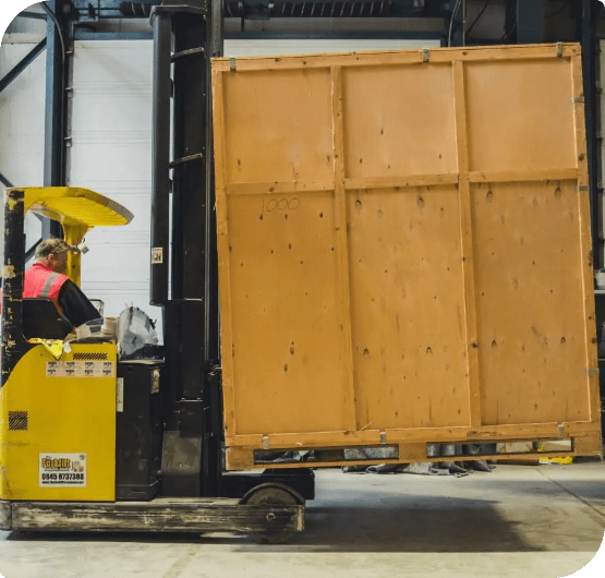 Worker operating a forklift carrying a large wooden crate in a warehouse.