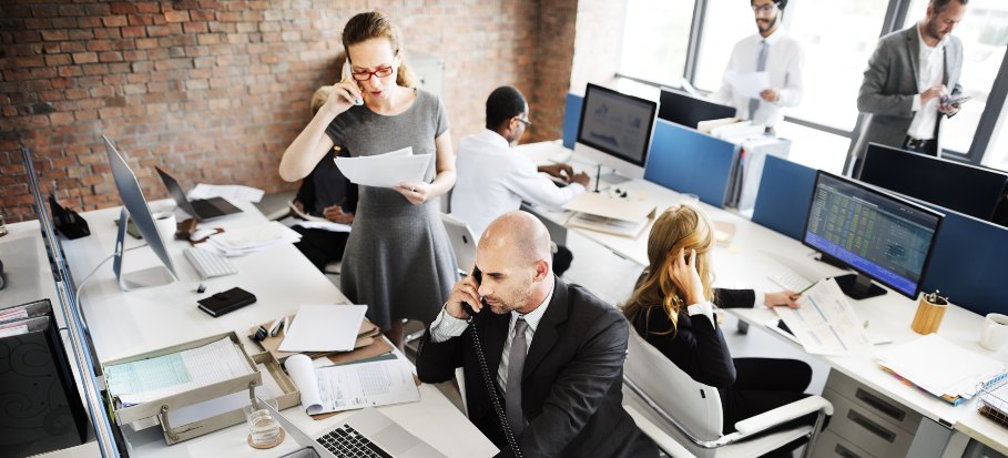 Busy office scene with multiple workers at desks with computers, papers, and phones.