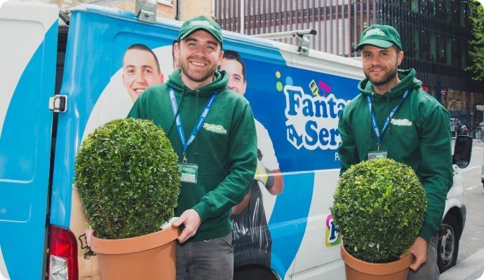 Two workers in green uniforms carrying potted plants beside a branded service van.