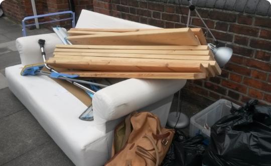 A pile of discarded items including a white sofa, wooden planks, a lamp and black bags on the street.